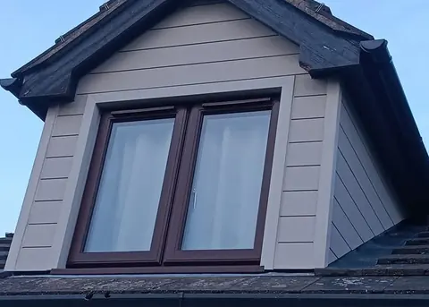 A dormer clad in stone-coloured boards around a rosewood window, on a tiled roof